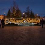 Happy Holidays script light display on a brick town plaza at dusk with lit evergreens and a holiday market behind it
